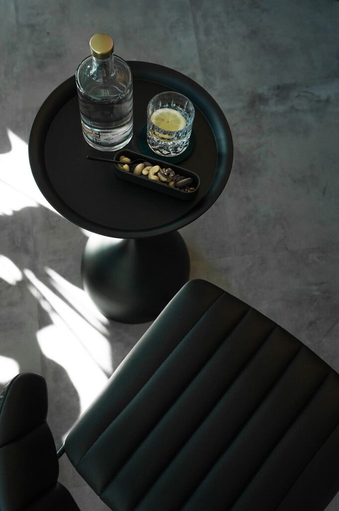 Stillleben mit Wasserkaraffe, Glas mit Zitronenscheibe und Snackschale auf schwarzem Beistelltisch neben gepolstertem Ledersitz.