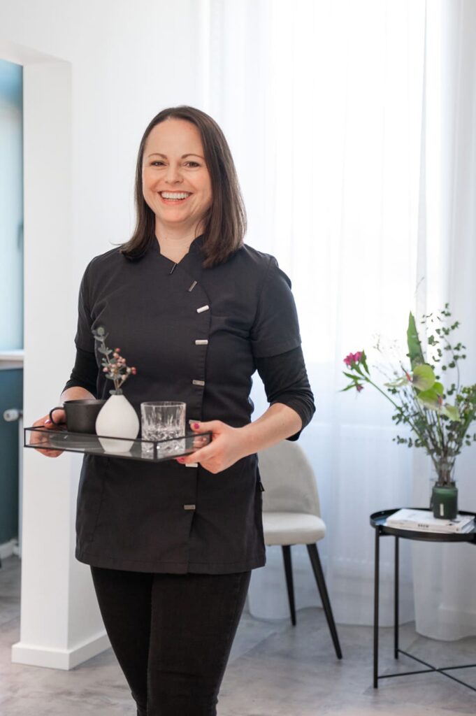 Claudia Behrmann, House Managerin, trägt Tablett mit Glas, Tasse und Vase, herzlich lächelnd in stilvollem, lichtdurchflutetem Raum.