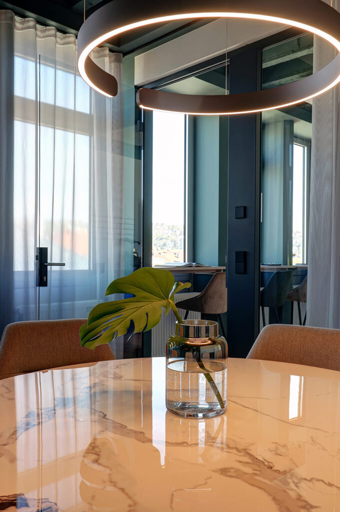 Marmortisch mit Glasvase und Monstera-Blatt vor Fensterfront und modernen Barhockern in stilvollem Meetingraum.