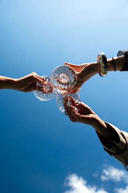 Three people raising glasses for a toast under a clear blue summer sky, capturing a social outdoor celebration.